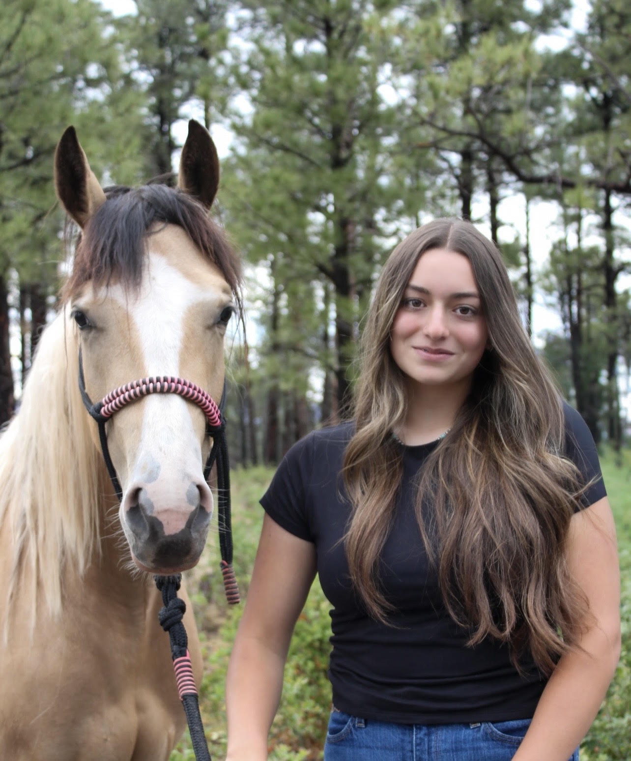 Sofia Garcia standing beside a buckskin horse in a forested New Mexico pasture