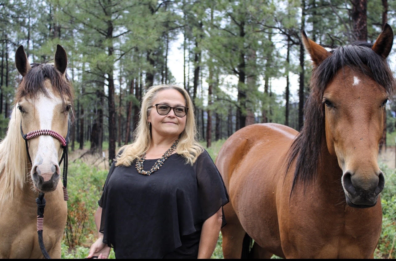Donna Abreu standing between two horses in a forested New Mexico pasture
