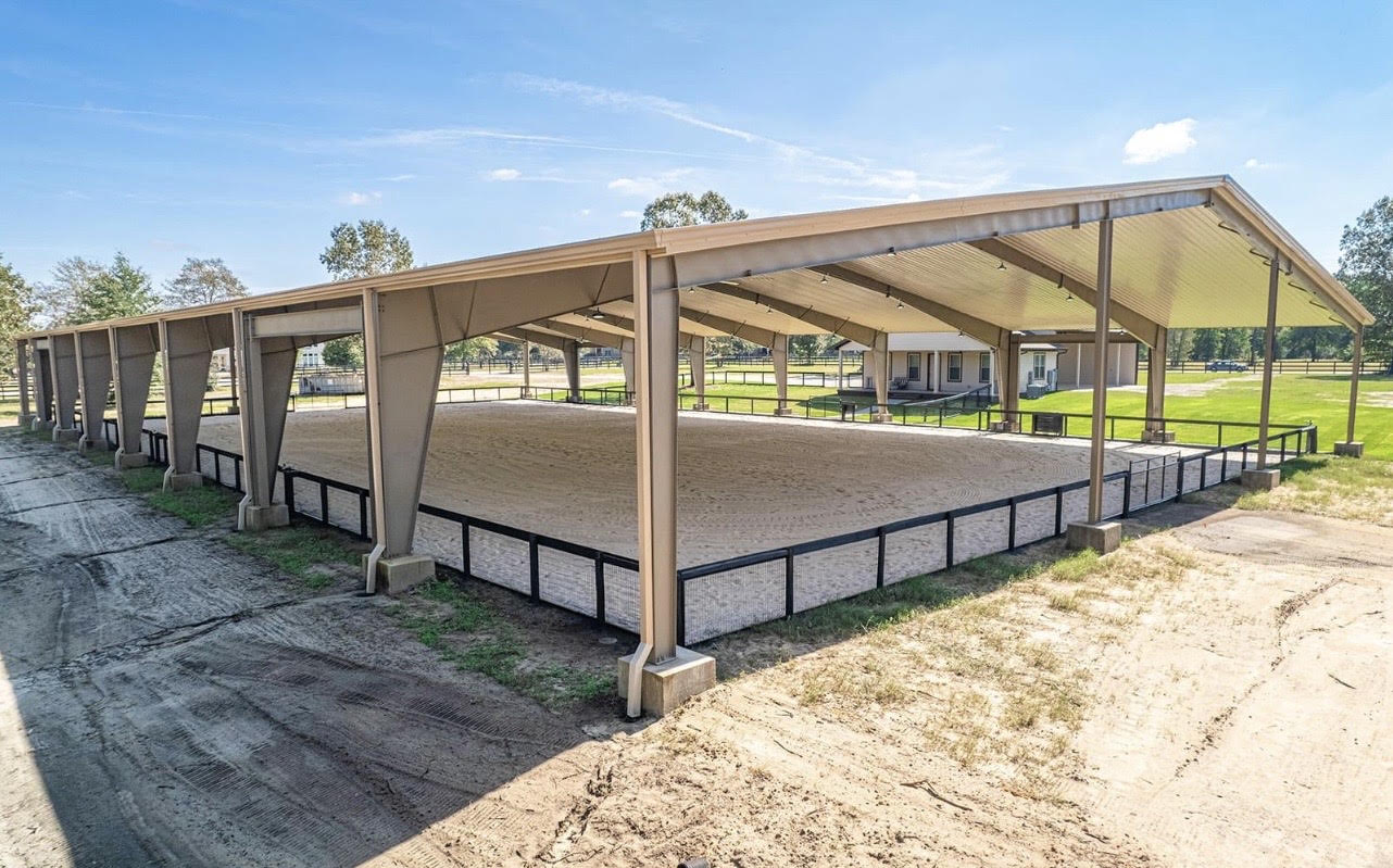 A covered equine arena with a metal roof and open sides, providing year-round protection from weather