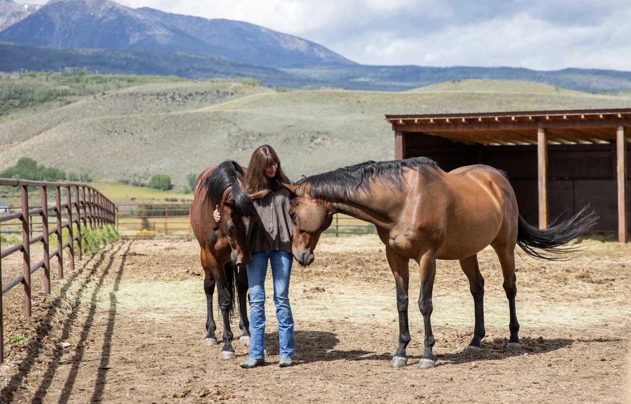 A woman standing between two horses in a peaceful ranch setting with mountains in the background